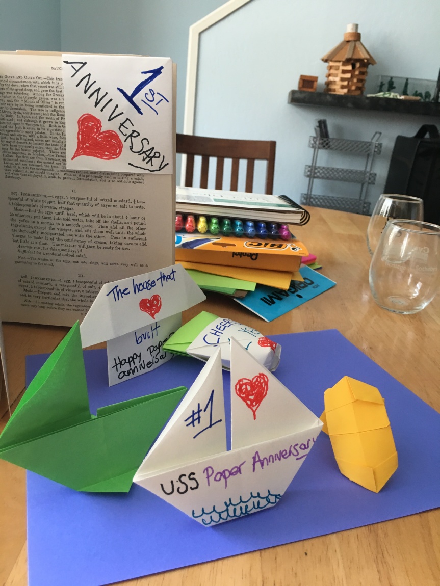 In the background, a book stands upright with a triangular bookmark attached to the top corner of a page, next to it, a stack of art supplies—spiral bound construction paper, a packet of markers, and a variety of colored papers, to the right of that stand two empty stemless wine glasses. In the foreground, origami sailboats, a house, cups, and a yellow ring, most items colored with hearts and messages of 1st anniversary celebration.