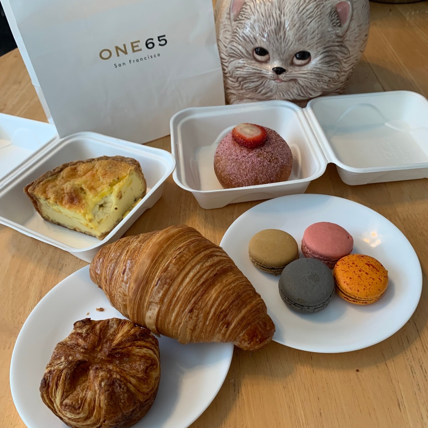 A white paper bag bearing the simple ONE65 logo stands next to a vintage cat faced cookie jar. In front are presented a collection of baked goods and pastries: A tall, firm wedge of pale quiche and a brioche roll topped with a round cross section of strawberry, each in their paper take out boxes, a large, golden, football-shaped croissant and a bundle of puff pastry known as the kouign amann, it's four corners turned in toward the center, on a small plate, and the four brightly colored macarons on another plate arranged in a diamond shape, gray, tan, orange with speckles, and pink.